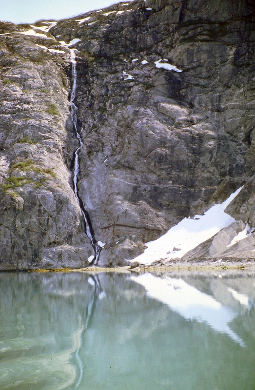 tall steep rocky cliff with long waterfall reflecting in calm waters in alaska
