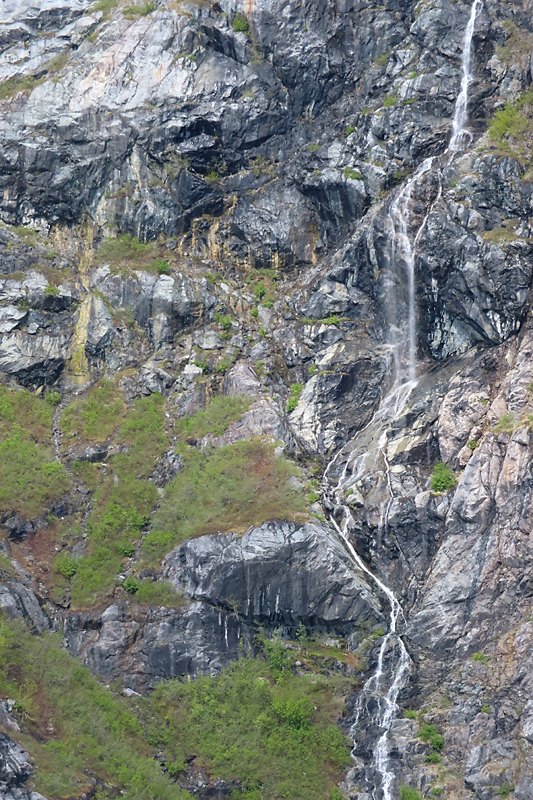 Long white narrow waterfall on grey cliffside with patches of green bushes in Alaska