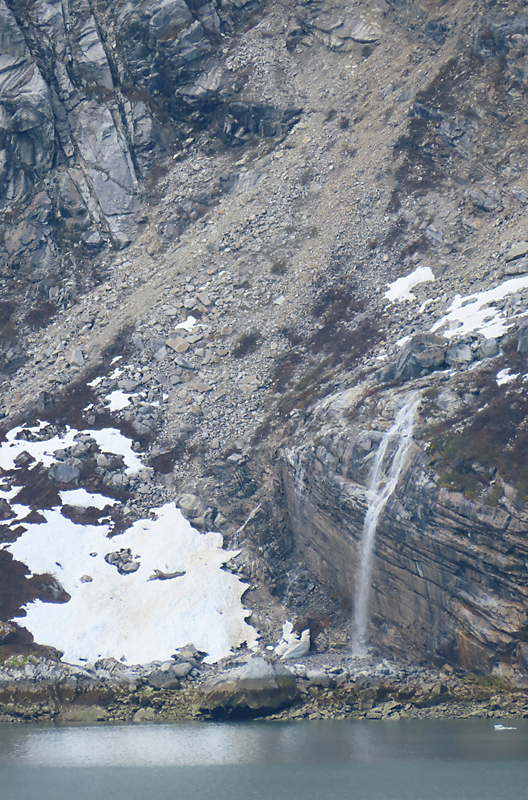 Waterfall in Alaska appears from grey cliffside and rocky terrain and snow patches